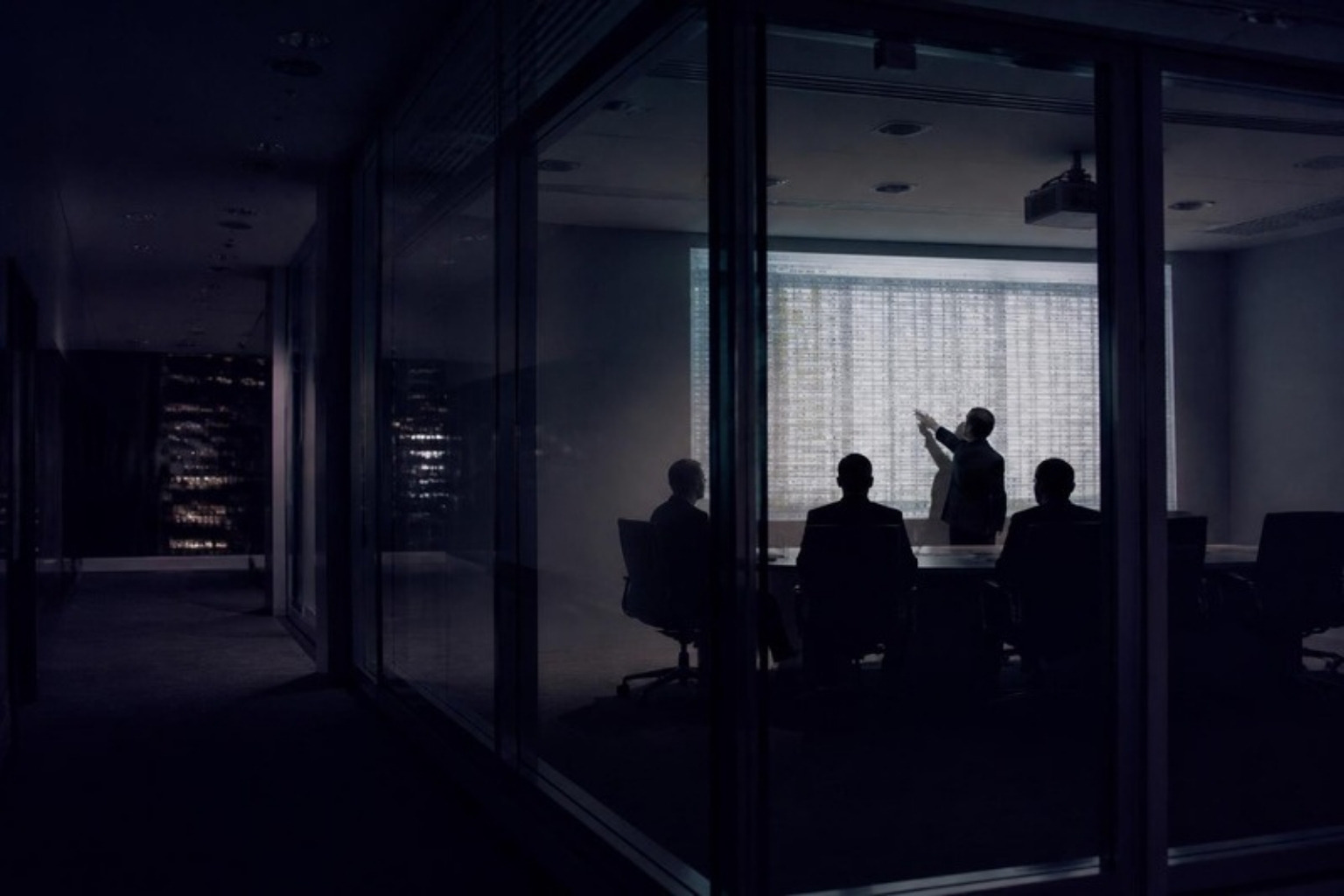 Executives in a dark boardroom, a projected spreadsheet grid on the wall