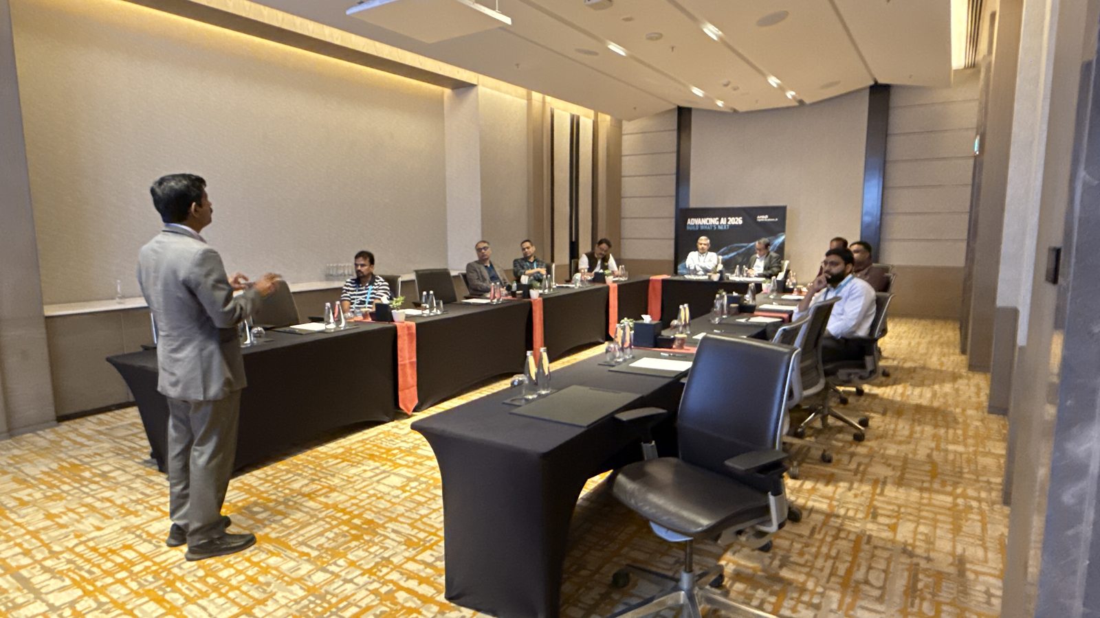 A speaker addresses technology leaders seated around a U-shaped table in a meeting room with an Advancing AI 2026 banner